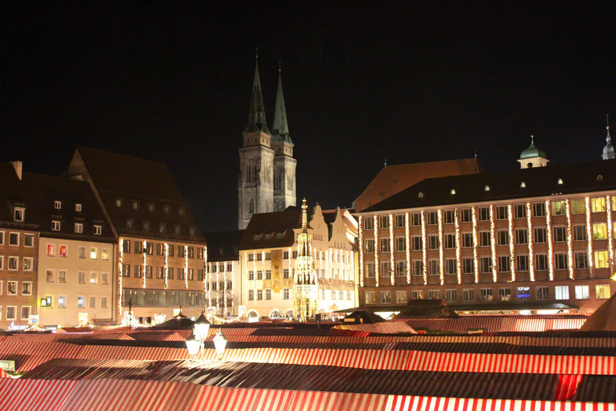 Toldos de rayas rojas y blancas en los puestos del Christkindlesmarkt de Núremberg de noche, con las dos torres de la iglesia de San Sebaldo detrás
