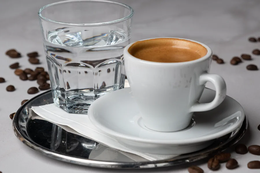Una taza de café expreso de cerámica blanca y un pequeño vaso de agua sobre una bandeja metálica