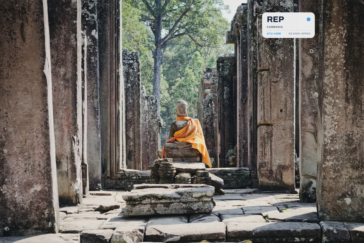 Una statua del Buddha in veste arancione tra i pilastri di pietra del tempio Bayon, con una scheda eSIM Atli che mostra il codice REP