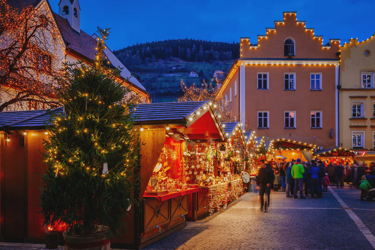 Der Weihnachtsmarkt auf dem Platz einer Kleinstadt in der Abenddämmerung mit beleuchteten Holzbuden und einem Weihnachtsbaum, dazwischen gehen Menschen