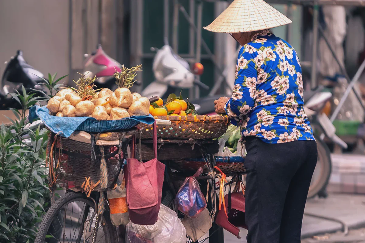 Una venditrice con cappello conico e una bicicletta carica di frutta in una strada vietnamita