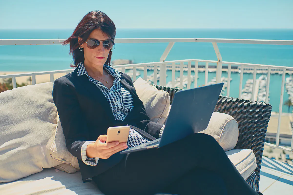 Una donna con il telefono in mano e il portatile aperto su una terrazza sopra il porto turistico