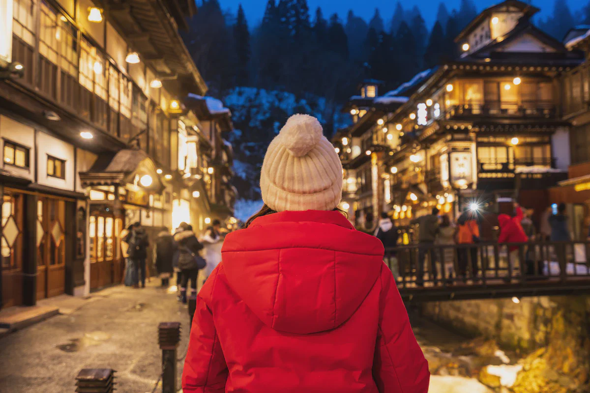 A traveller in a red jacket looking along the lit wooden inns of Ginzan Onsen on a snowy evening
