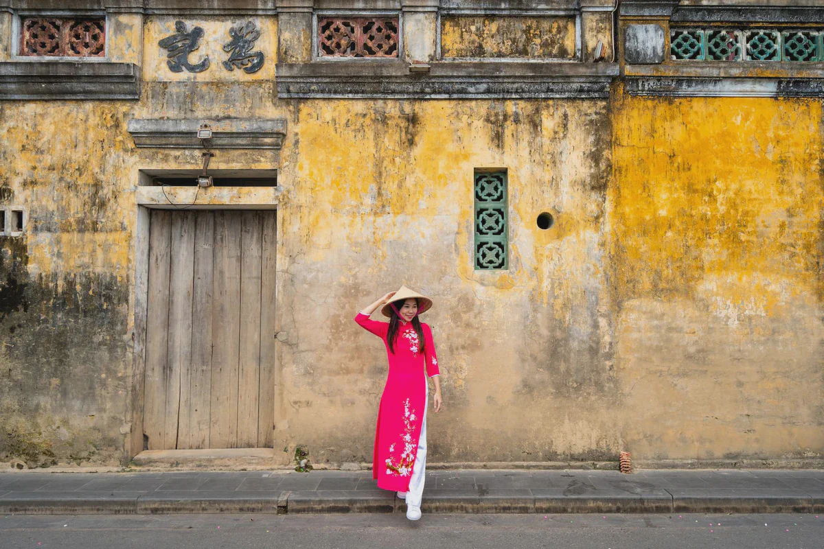 Una donna in lungo abito rosa e cappello conico accanto a un muro giallo consumato a Hoi An