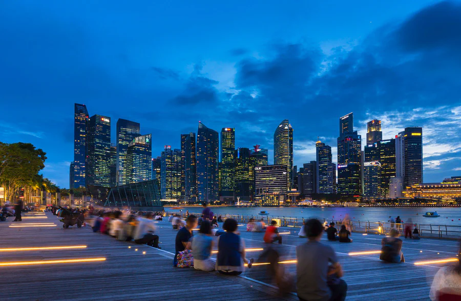People sitting on a wooden waterfront promenade at dusk, looking across the bay at the Singapore skyline